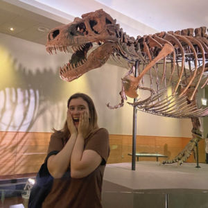 Lucy stands in front of a skeletal model of a T-Rex. Her hands are on her face in fake shock and she smiles at the camera.